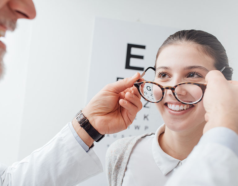 A imagem mostra uma mulher branca, de cabelos presos, dando um largo sorriso enquanto um médico de barba branca coloca um óculos de grau nos olhos dela. Ao fundo, vê-se um cartaz de teste de visão, com letras grandes e pequenas.